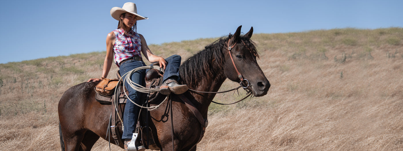 Cowgirl sitting on a horse in a field wearing a hat, plaid shirt, and jeans