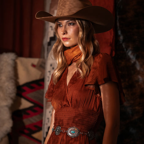A woman wearing a brown cowboy hat, rust-orange dress, and silver concho belt, posed in a warm, dimly lit rustic interior.