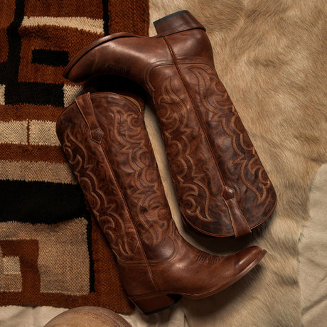 A pair of brown leather cowboy boots with intricate hand-tooled western stitching, displayed on a patterned rug and a beige fur surface.