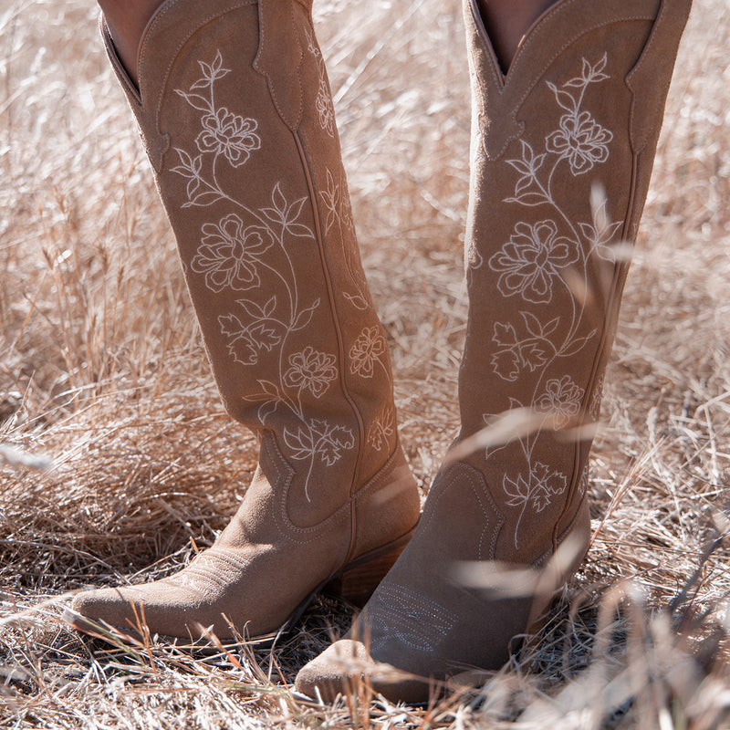 A close-up of a person's legs wearing tan suede cowboy boots with white floral embroidery, standing in dry, golden grass.