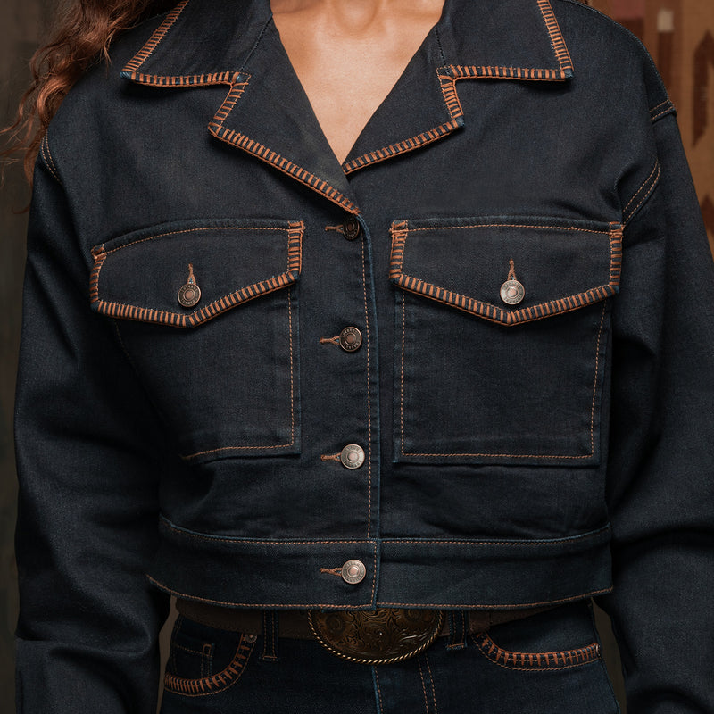 A cropped dark indigo denim jacket with contrasting orange-brown contrast stitching and silver-toned metal snap buttons on the chest pockets, shown on a model.