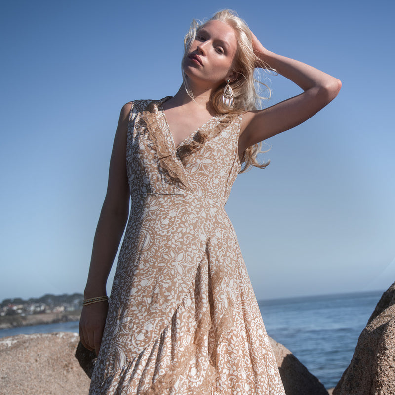 A woman wearing a sleeveless beige and white patterned dress stands outdoors by the ocean, with rocks and blue sky in the background.
