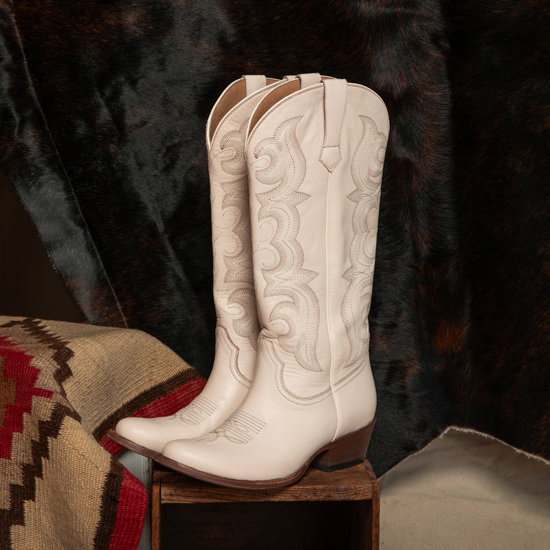 A pair of off-white western cowboy boots with intricate embroidery, displayed on a wooden block against a dark brown fur backdrop and a patterned textile.