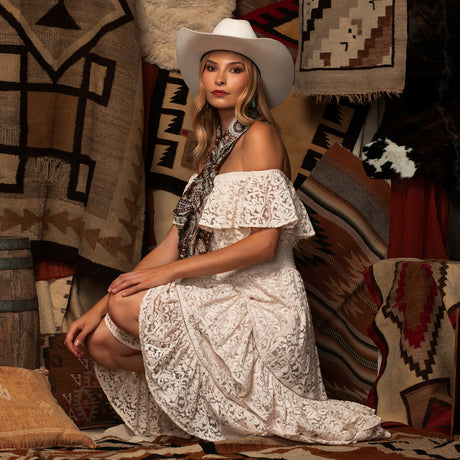 A woman wearing a white off-the-shoulder lace maxi dress, a patterned scarf, and a white cowboy hat sits on a patterned rug surrounded by southwestern-style woven textiles.