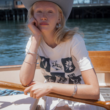 A woman wearing a white graphic t-shirt and a wide-brimmed gray hat sits on a wooden boat with water in the background.