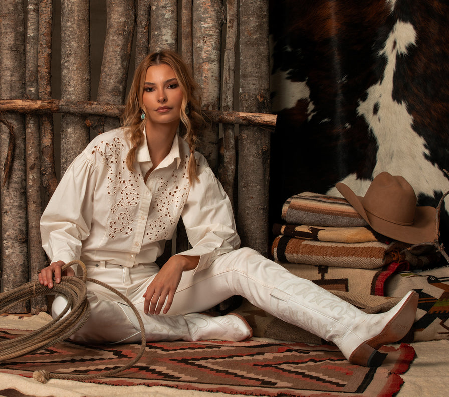 A woman sits on a patterned rug in a rustic western-themed setting, wearing a matching cream-colored embroidered shirt and pants with white cowboy boots. She holds a lasso, with a brown hat, folded blankets, and a cowhide backdrop behind her.